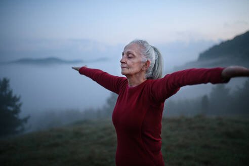 A senior woman doing breathing exercise in nature on early morning with fog and mountains in background. - HPIF01474