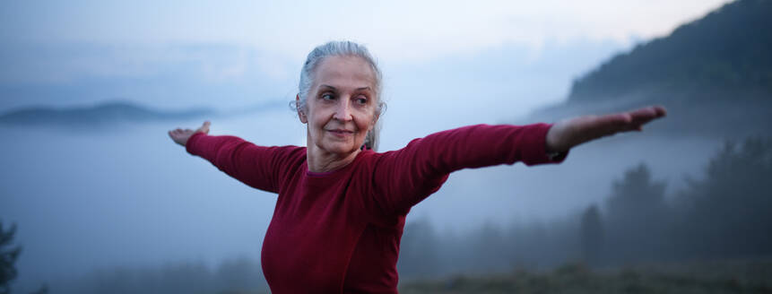 A senior woman doing breathing exercise in nature on early morning with fog and mountains in background. - HPIF01475
