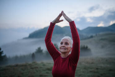 Eine ältere Frau macht Atemübungen in der Natur am frühen Morgen mit Nebel und Bergen im Hintergrund. - HPIF01476