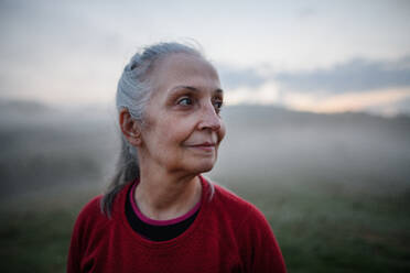 Eine fröhliche ältere Frau in der Natur am frühen Morgen mit Nebel und Bergen im Hintergrund. - HPIF01482