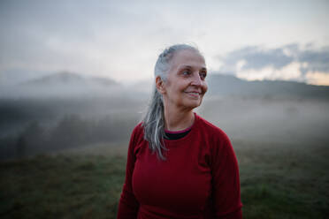 Eine fröhliche ältere Frau in der Natur am frühen Morgen mit Nebel und Bergen im Hintergrund. - HPIF01483