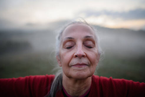 A senior woman doing breathing exercise in nature on early morning with fog and mountains in background, close-up - HPIF01485