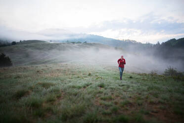 Eine ältere Frau joggt in der Natur am frühen Morgen mit Nebel und Bergen im Hintergrund. - HPIF01489