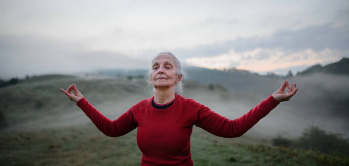 A senior woman doing breathing exercise in nature on early morning with fog and mountains in background. - HPIF01492
