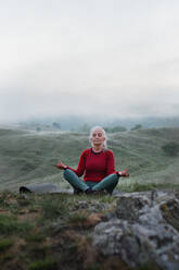 Eine ältere Frau macht Atemübungen in der Natur am frühen Morgen mit Nebel und Bergen im Hintergrund. - HPIF01495