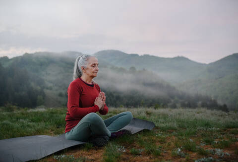 A senior woman doing breathing exercise in nature on early morning with fog and mountains in background. - HPIF01497