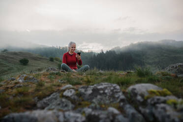 Eine ältere Frau, die ein Selfie macht, während sie eine Atemübung in der Natur am frühen Morgen mit Nebel und Bergen im Hintergrund macht. - HPIF01498