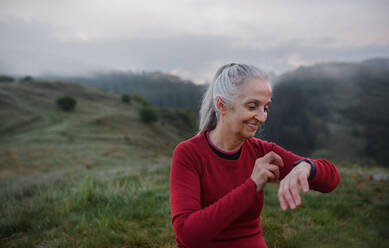 A senior woman jogger setting and looking at sports smartwatch, checking her performance in nature on early morning with fog and mountains in background. - HPIF01500