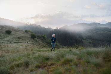 Eine ältere Frau joggt in der Natur am frühen Morgen mit Nebel und Bergen im Hintergrund. - HPIF01506