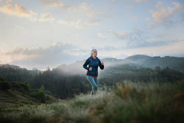 A senior woman jogging in nature on early morning with fog and mountains in background. - HPIF01507