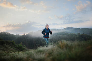 Eine ältere Frau joggt in der Natur am frühen Morgen mit Nebel und Bergen im Hintergrund. - HPIF01507