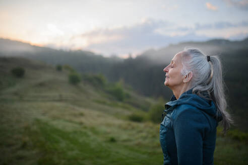 A senior woman doing breathing exercise in nature on early morning with fog and mountains in background. - HPIF01509