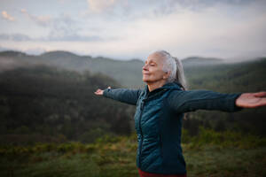 Eine ältere Frau macht Atemübungen in der Natur am frühen Morgen mit Nebel und Bergen im Hintergrund. - HPIF01511