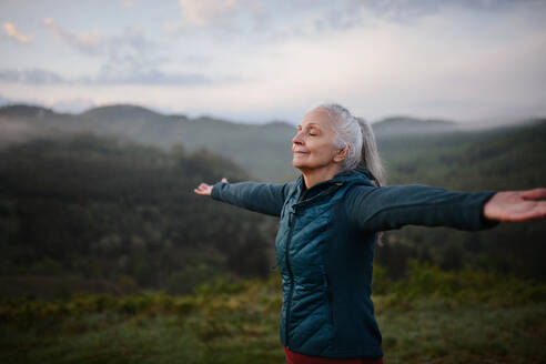 A senior woman doing breathing exercise in nature on early morning with fog and mountains in background. - HPIF01511