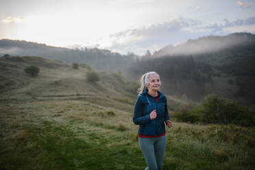 Eine ältere Frau joggt in der Natur am frühen Morgen mit Nebel und Bergen im Hintergrund. - HPIF01514