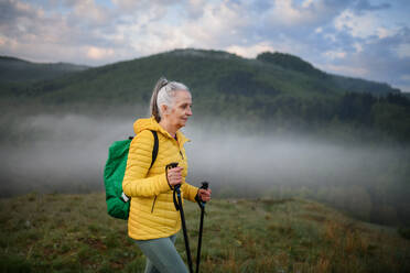 Eine ältere Frau beim Wandern in der Natur am frühen Morgen mit Nebel und Bergen im Hintergrund. - HPIF01518