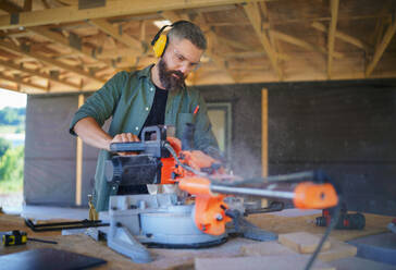 Bauarbeiter arbeitet mit elektrischer Säge in einer Holzkonstruktion eines Hauses, diy umweltfreundliche Häuser Konzept. - HPIF01526