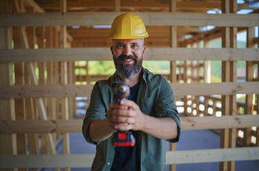 Construction worker posing with electric screwdriver in a wooden frame building, diy eco-friendly homes concept. - HPIF01558
