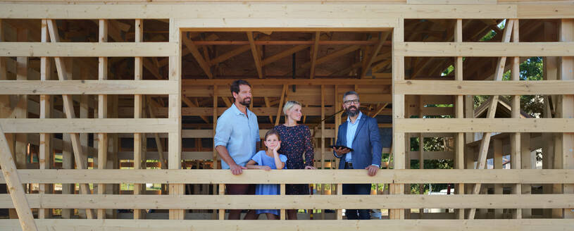 Ein Handelsvertreter zeigt einer jungen Familie ein unfertiges ökologisches Holzhaus auf der Baustelle und betrachtet gemeinsam die Aussicht. - HPIF01567