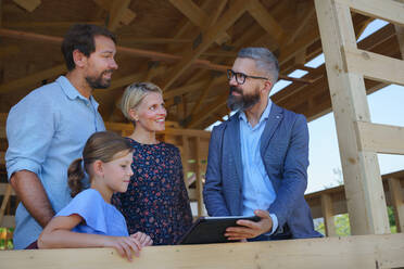 Ein Handelsvertreter zeigt einer jungen Familie auf der Baustelle die Pläne eines neuen unfertigen ökologischen Holzhauses und betrachtet gemeinsam die Aussicht. - HPIF01572