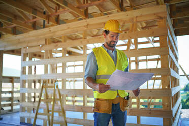 Construction engineer or architect with blueprints visiting and chechking eco building site of a wood frame house. - HPIF01589