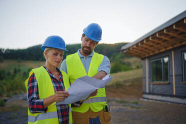 Bauingenieure oder Architekten prüfen einen Bauplan vor einem unfertigen Öko-Rahmenhaus. - HPIF01601