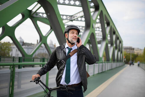 A portrait of businessman commuter on the way to work, pushing bike and calling on mobile phone, sustainable lifestyle concept. - HPIF01620