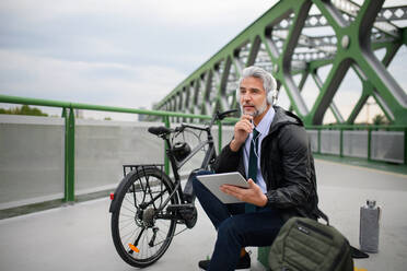 Ein Geschäftsmann mit Fahrrad, der auf einer Brücke sitzt, ein Tablet benutzt und Musik hört. Pendeln und alternatives Verkehrskonzept - HPIF01621