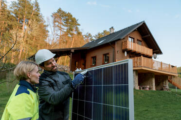 Ein lächelnder Handwerker, der ein Solarmodul trägt, während er ein Solarmodulsystem auf einem Haus installiert. - HPIF01663