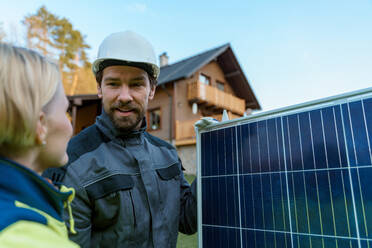 Ein lächelnder Handwerker, der ein Solarmodul trägt, während er ein Solarmodulsystem auf einem Haus installiert. - HPIF01664