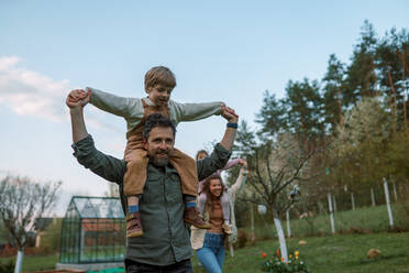 Glückliche Eltern mit kleinen Kindern auf Huckepack, die gemeinsam in einem Garten in der Nähe ihres Hauses rennen und Spaß haben. - HPIF01672