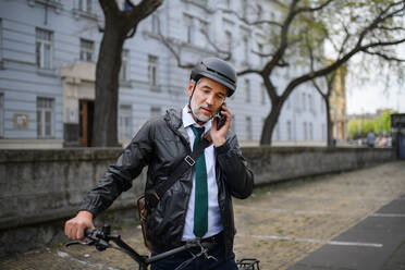 Porträt eines Geschäftsmannes auf dem Weg zur Arbeit, der sein Fahrrad schiebt und mit seinem Handy telefoniert, ein Konzept für nachhaltigen Lebensstil. - HPIF01764