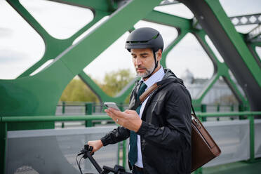Ein Geschäftsmann auf dem Weg zur Arbeit, der sein Fahrrad auf der Brücke schiebt und eine SMS auf seinem Handy schreibt, ein nachhaltiges Lebensstilkonzept. - HPIF01767