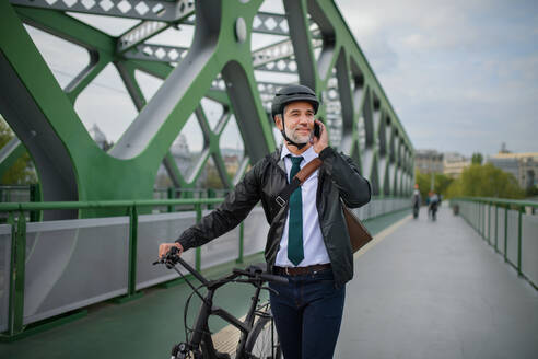 Ein Geschäftsmann auf dem Weg zur Arbeit, der sein Fahrrad auf der Brücke schiebt und mit dem Handy telefoniert, ein nachhaltiges Lebensstilkonzept. - HPIF01768