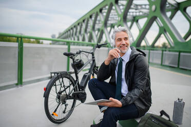 Ein Geschäftsmann mit Fahrrad, der auf einer Brücke sitzt, ein Tablet benutzt und Musik hört. Pendeln und alternatives Verkehrskonzept - HPIF01779