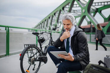 Ein Geschäftsmann mit Fahrrad, der auf einer Brücke sitzt, ein Tablet benutzt und Musik hört. Pendeln und alternatives Verkehrskonzept - HPIF01780