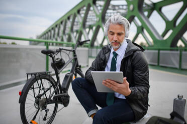 Ein Geschäftsmann mit Fahrrad, der auf einer Brücke sitzt, ein Tablet benutzt und Musik hört. Pendeln und alternatives Verkehrskonzept - HPIF01781