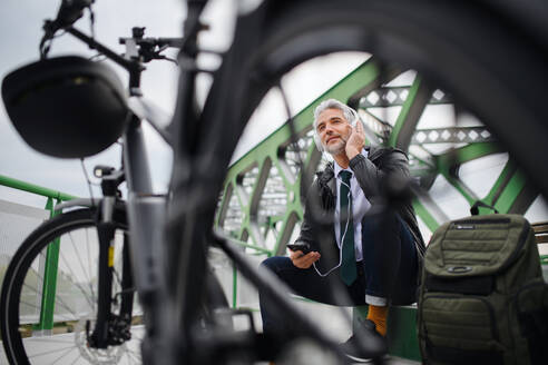 Ein Geschäftsmann mit Fahrrad, der auf einer Bank sitzt und ein Smartphone benutzt. Pendeln und alternatives Verkehrskonzept - HPIF01785