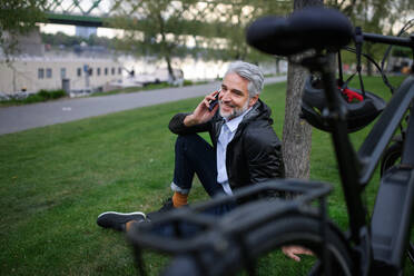 A businessman with bike sitting on grass in park, using smartphone. Commuting and alternative transport concept - HPIF01799