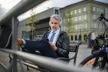 A businessman with bike sitting on bench, listening to music with feet up and resting. Commuting and alternative transport concept - HPIF01804