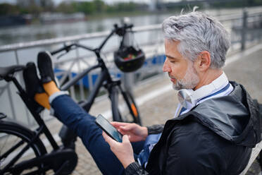 Ein Geschäftsmann mit Fahrrad, der auf einer Bank sitzt und ein Smartphone benutzt. Pendeln und alternatives Verkehrskonzept - HPIF01806