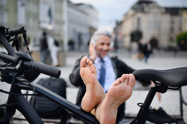 Ein Geschäftsmann mit Fahrrad auf einer Bank sitzend, Musik hörend, die Füße hochgelegt und sich ausruhend. Pendeln und alternatives Verkehrskonzept - HPIF01809
