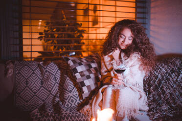 A happy young woman wrapped in blanket sitting on terrace and drinking wine in the evening. - HPIF01820