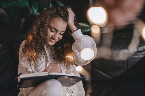 Eine glückliche junge Frau sitzt auf dem Sofa und schreibt in Tagebuch am Abend in gemütlichen hyyge Wohnzimmer. - HPIF01823
