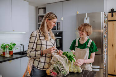 A mother unpacking local food in zero waste packaging from bag with help of daughter in kitchen at home. - HPIF01839