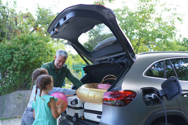 Eine Familie mit kleinen Kindern, die ihr Auto belädt und darauf wartet, dass das Auto aufgeladen wird, bevor sie zum Picknick aufbricht. - HPIF01862