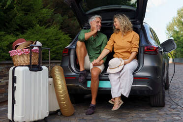 Ein Ehepaar mittleren Alters sitzt im Kofferraum und wartet darauf, dass sein Auto aufgeladen wird, bevor es in den Sommerurlaub fährt. - HPIF01876