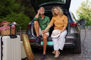 Ein Ehepaar mittleren Alters sitzt im Kofferraum und wartet darauf, dass sein Auto aufgeladen wird, bevor es in den Sommerurlaub fährt. - HPIF01877