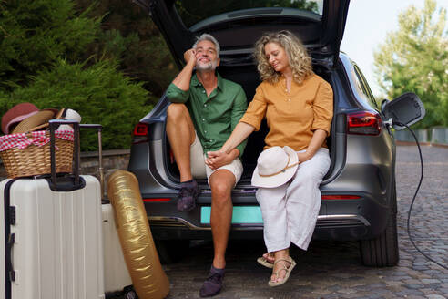 A middle-aged couple sitting in trunk while waiting for charging car before travelling on summer holiday. - HPIF01877