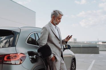 Ein Geschäftsmann hält sein Smartphone in der Hand, während er sein Auto an einer Ladestation für Elektrofahrzeuge auflädt, Nahaufnahme. - HPIF01900